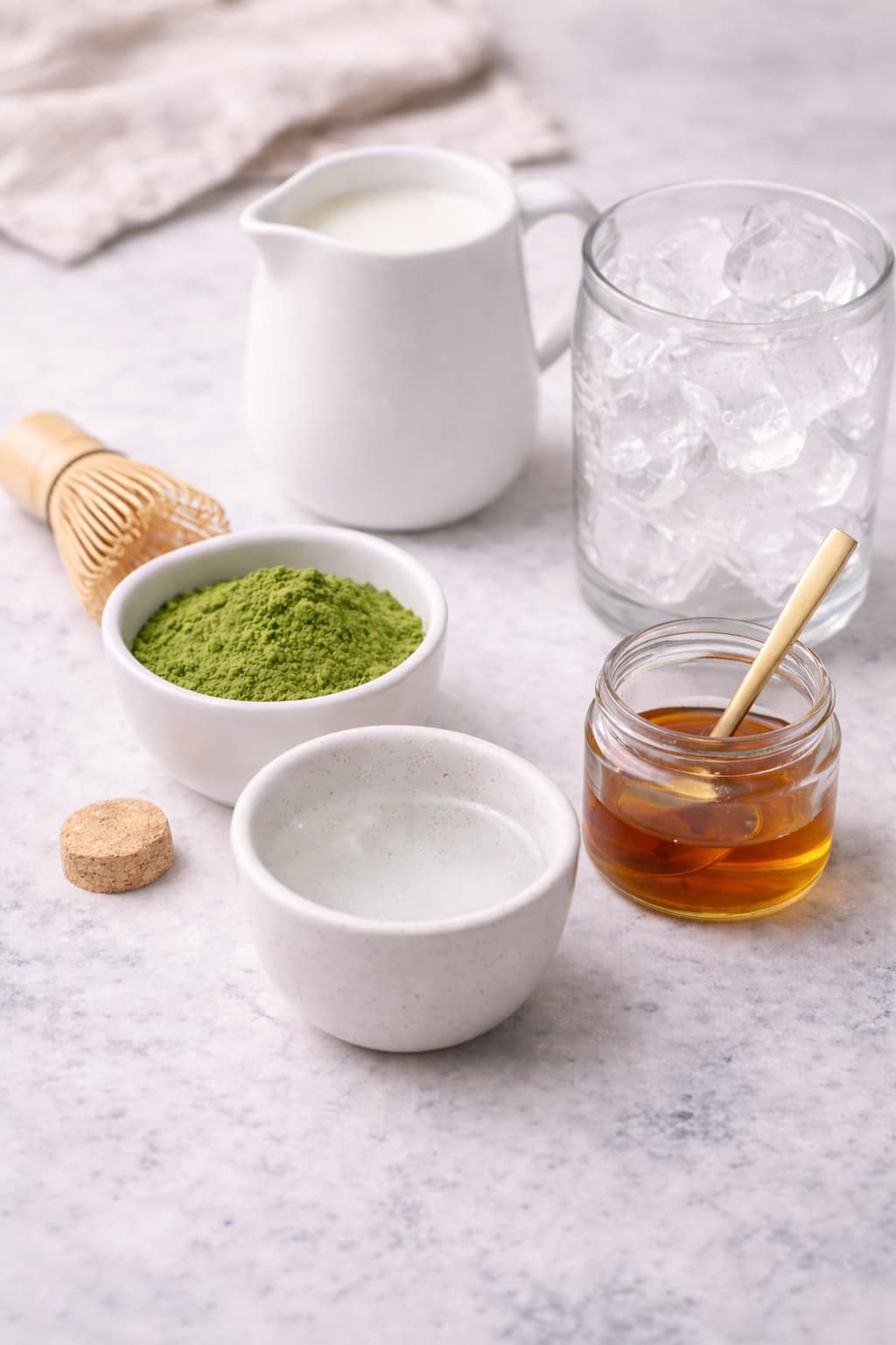 Ingredients for an iced matcha latte including ceremonial-grade matcha powder, milk, ice, sweetener, and water